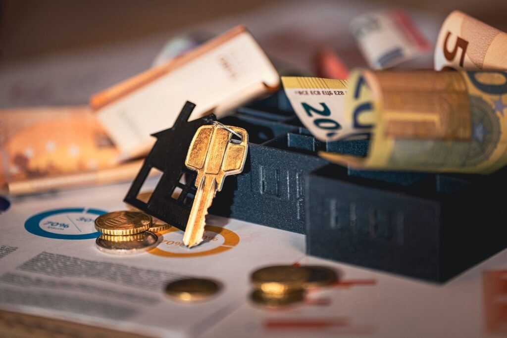 Llave con llavero en forma de casa junto a maquetas de edificios, billetes y monedas de euro sobre unos documentos