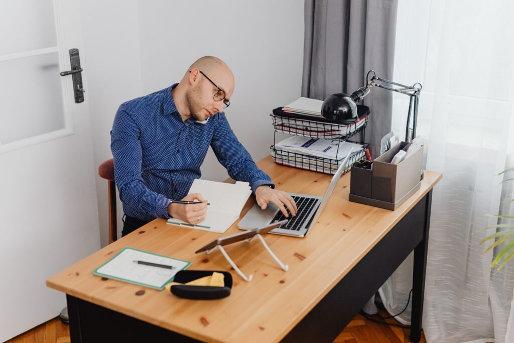 Hombre teletrabajando desde casa con un portátil mientras habla por teléfono y toma notas